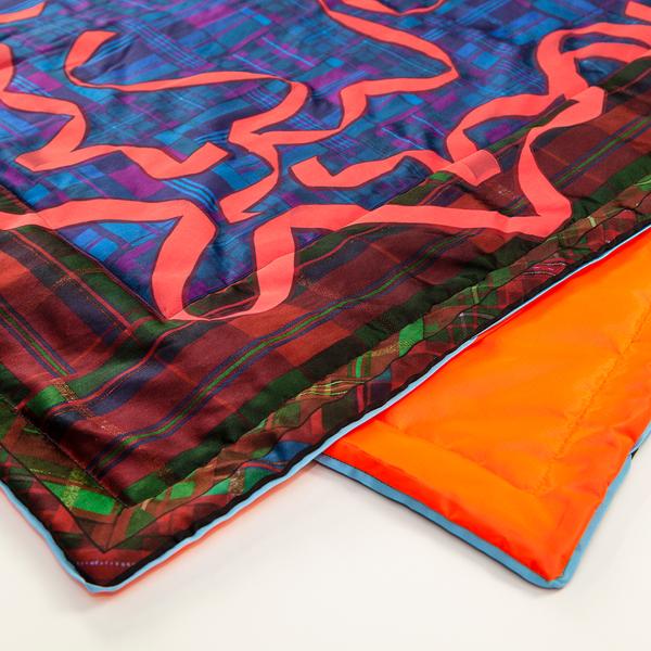 Two offset corners of a folded blanket show the top layer, with a plaid border and bright red ribbons over a deep blue ground, and its orange underside.