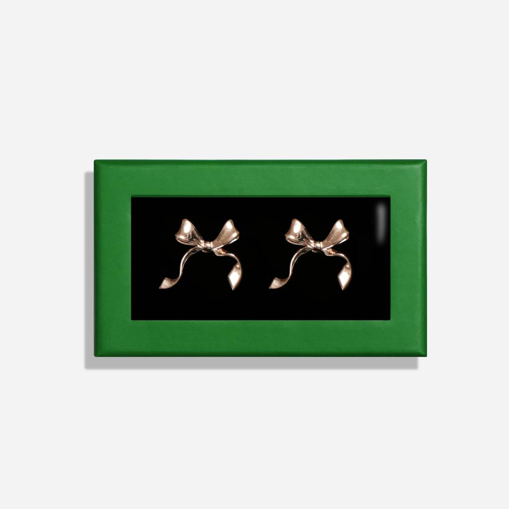 A rectangular box with a green border features a pair of rose gold colored cuff links with a decorative bow inside.