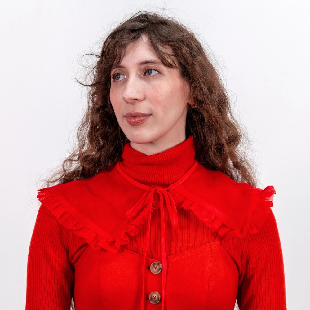 A photo of a model wearing a red silk organza removable collar around her neck and sitting atop her shoulders. The collar has a rounded edge and ruffle going all the way around. The tie is silk and ties in a bow.