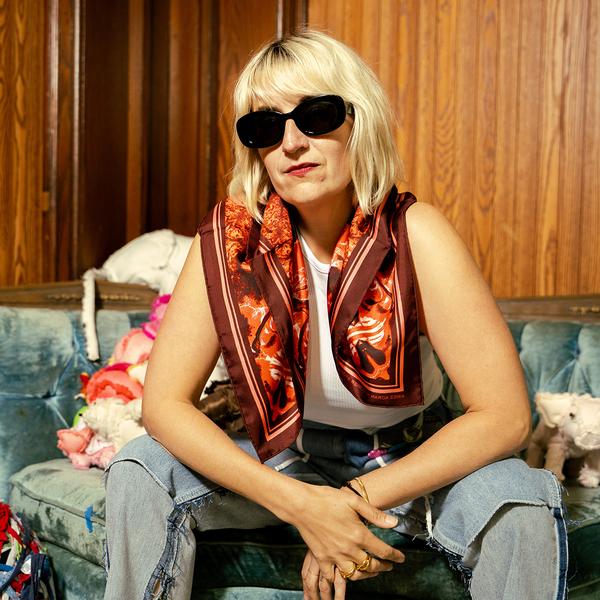 The artist Aimee Koran, a blonde woman, models her red scarf while seated on a couch. She is wearing sunglasses and the scarf loose around her shoulders. The scarf has a maroon border surrounding abstract red textures.