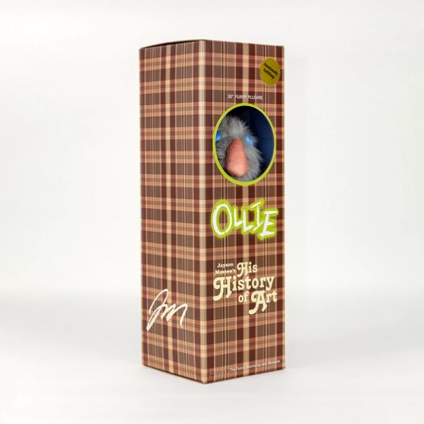 A photo of a tall brown plaid box in which two of its sides are visible. On the lower left is the artist Jayson Musson's signature. The right side reads "Ollie" and "Jayson Musson's His History of Art" and has a circular viewing window that reveals a gray stuffed animal inside.