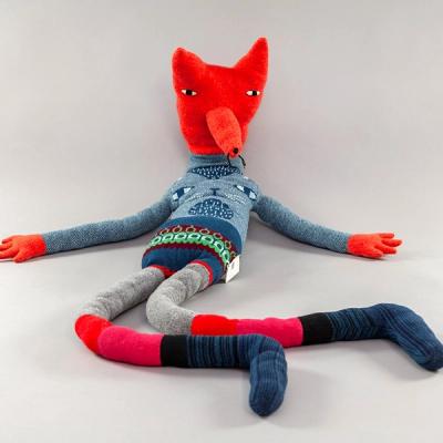 A large plush with very long tube-like limbs laid out with its red fox-like head sitting upright. Its small eyes are very far apart and sit well above its protruding snout. It is wearing a grey sweater with stitching that resembles the face of another animal, possibly a cat. Its legs feature various bands of gray, red, and pink with dark blue and black striped socks.
