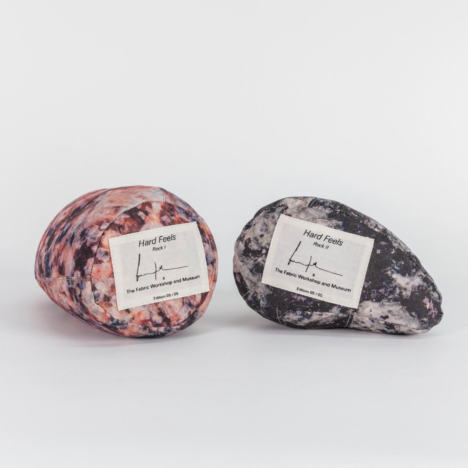 A photograph of the bottom sides of two small soft sculptures covered with patterns that resemble polished stone of different hues. Each one has a label reading the title of the edition (Hard Feels), the artist's signature, and edition number.