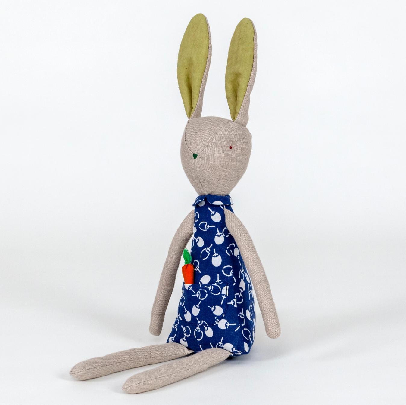 A photograph of a plush rabbit made of light beige woven fabric characterized to have a pointy nose, triangular shaped body, and long thin arms and legs. The body of the rabbit is made of a primarily blue patterned fabric and includes a pocket with a removable plush orange carrot.