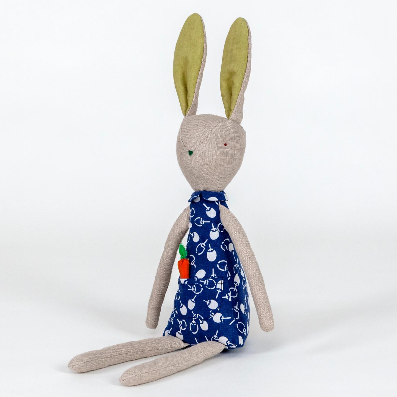 A photograph of a plush rabbit made of light beige woven fabric characterized to have a pointy nose, triangular shaped body, and long thin arms and legs. The body of the rabbit is made of a primarily blue patterned fabric and includes a pocket with a removable plush orange carrot.