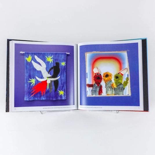 A book spread featuring two images (one on each page) of installation photos from the exhibition Jayson Musson: His History of Art. On the left is a window curtain referencing Gentileschi's "Judith Slaying Holofernes" in the style of Matisse's "Icarus." On the right is an image of three puppets wearing denim vests and pope hats.