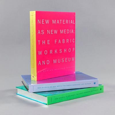 A photograph of a magenta book with a yellow spine stacked on two other books of the same size but in a light blue and green. The book reads "New Material as New Media: The Fabric Workshop and Museum."