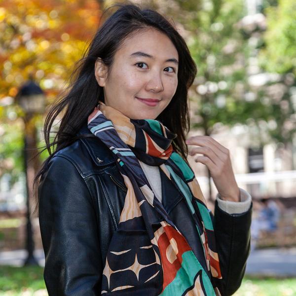 A person with straight dark hair is wearing the scarf over a leather jacket and smiling at the camera. Out of focus in the background, there is a city park with fall foliage.