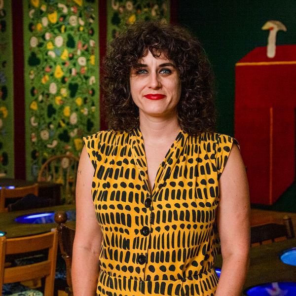 A portrait of the artist Jessica Campbell, a white woman with curly brown hair. She is wearing a sleeveless yellow blouse with vertical black patterned markings. She is standing in a dimly lit room with green and red artworks in the background and a set of tables and chairs.