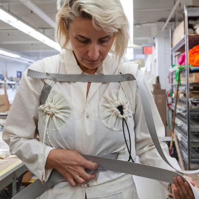 A photo of a white woman with blonde hair, the artist Aimee Koran, trying on a white jumpsuit with drawstrings positioned over her chest. She is stretching out a strap of fabric, presumably to tighten or measure it according to her body.