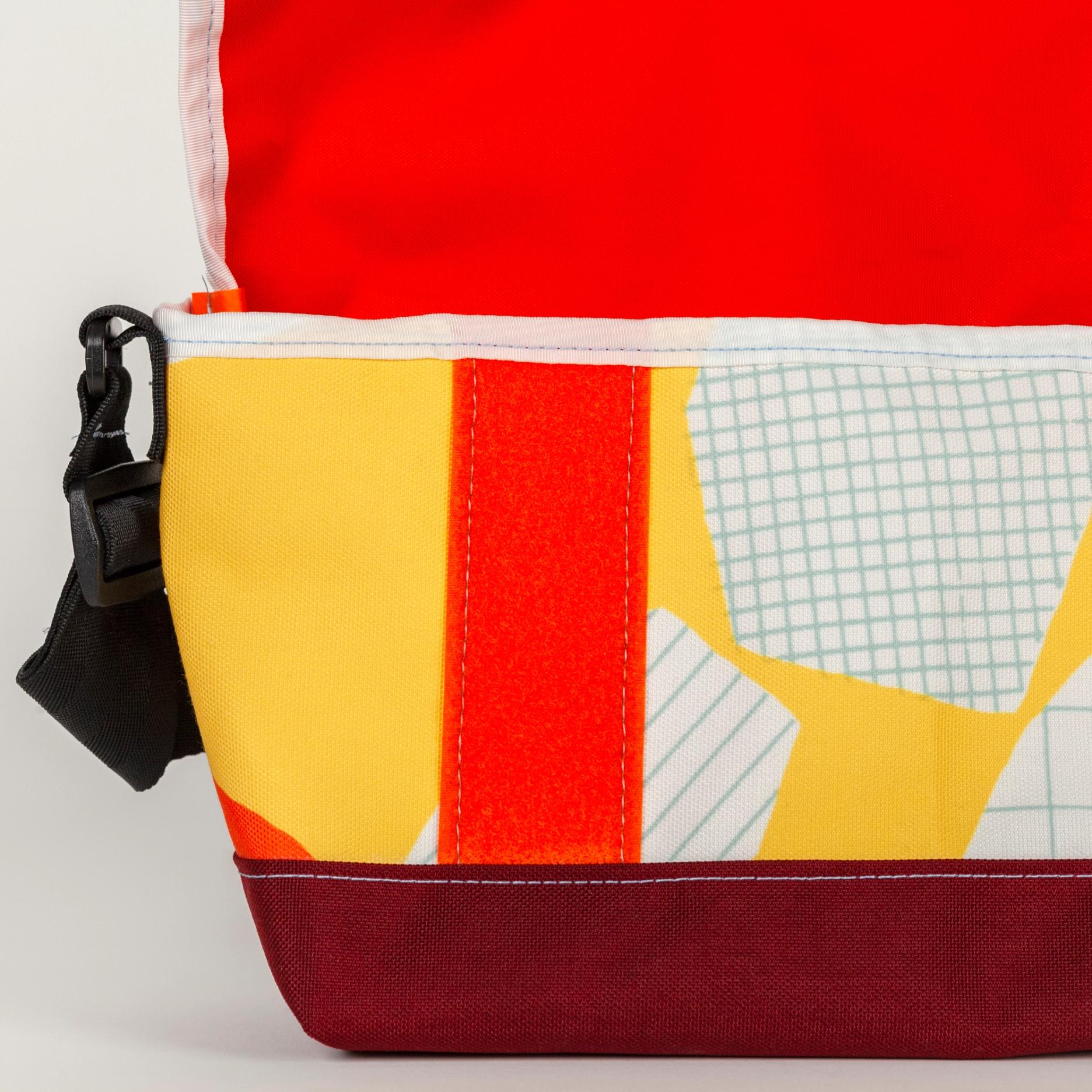 A detail shot of a shallow, wide, vibrant yellow bag with bold abstract shapes (bright orange, white with light blue stripes, and white with a light blue grid pattern). The top flap is open revealing a bright orange lining with a strip of orange velcro on the side of the bag. There is a white seam along the flap and opening of the bag. The bag has a black strap and the base of the bag is solid maroon.