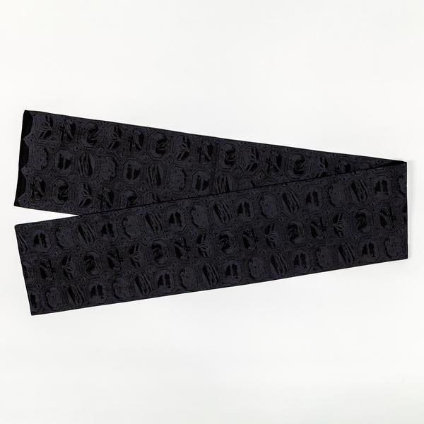A detail of a charcoal black scarf with a grid of octagonal vignettes also printed in black. Each features an abstract composition. Two ends of the scarf are folded over one another to form a V-shape.