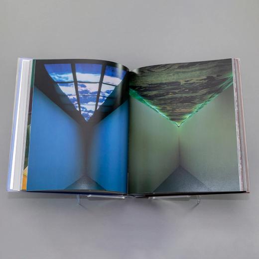 A photo of a book spread with a full-bleed photograph of artwork by Mel Chin on each page. On the left is a mostly blue picture of a corner of a room with a triangular grid of clouds above. On the right is a mostly green corner of a room with a wave-like texture on the triangular-shaped ceiling above. The edges of the wall and ceiling glow a bright green.