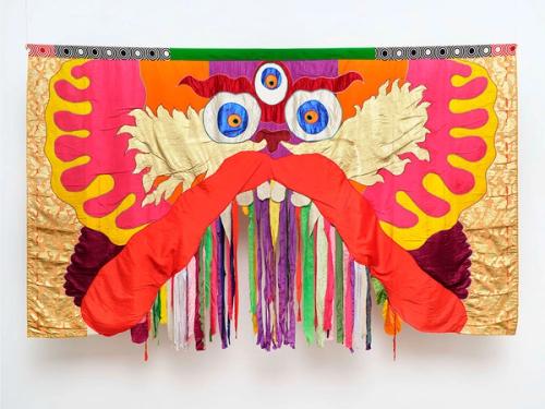 A photo of a fabric-based artwork of the face of a three-eyed dragon staring directly at the viewer. The face is cartoonish and mostly symmetrical, with different colors of fabric emanating outwardly from its nose. It has a large red upper lip that bows upward, with colorful tassels hanging under its teeth.