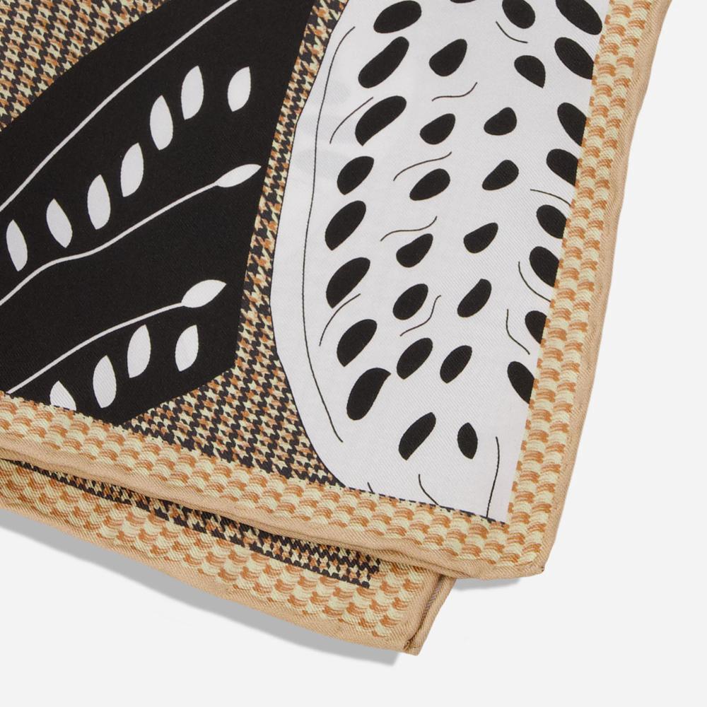 A folded pocket square with a detail of the corner. Its tan border resembles a woven texture while the rest shows a black and tan houndstooth pattern. Overtop is a printed black and white illustration of a house sparrow’s wings.