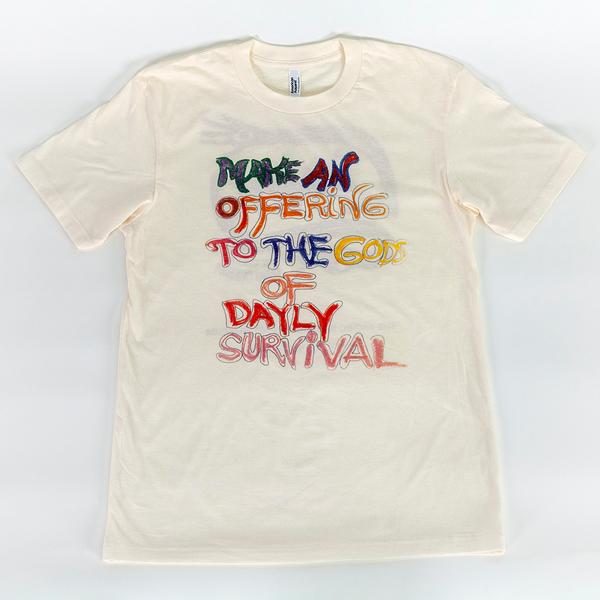 An off-white t-shirt that reads in a hand-drawn message, "Make An Offering to the Gods of Daily Survival." The word daily is spelled d-a-y-l-y. Each word is colored in with varying colors, including some individualized letters.