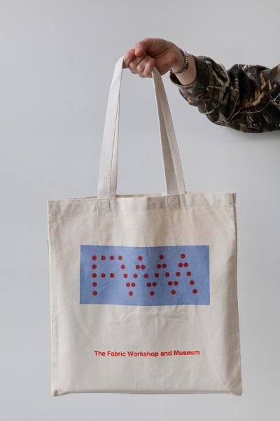 A photo of a hand model holding a light tan canvas tote bag. On the bag, there is a screenprinted light blue rectangle (landscape). Inside the light blue rectangle are the letters "FWM" and the letters are made up of bold, dark red dots. Underneath the rectangle in a smaller, bold, red font reads "The Fabric Workshop and Museum"