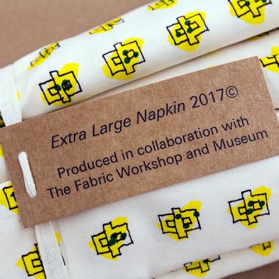 A detail of rolled napkins that show a repeated emblem printed with black lines and yellow. At the center of the image is a tag that says, "Extra Large Napkins 2017. Produced in collaboration with The Fabric Workshop and Museum."