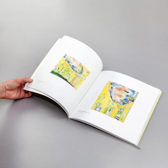 A photograph of a hand holding a book open. The two pages feature mostly yellow paintings of abstract interiors.