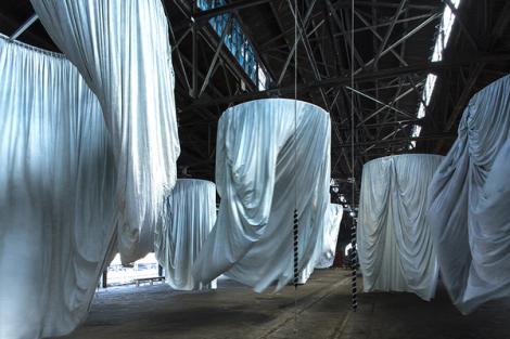 A photograph of a vast warehouse with a series of draped white curtains suspended in the form of large cylinders. 