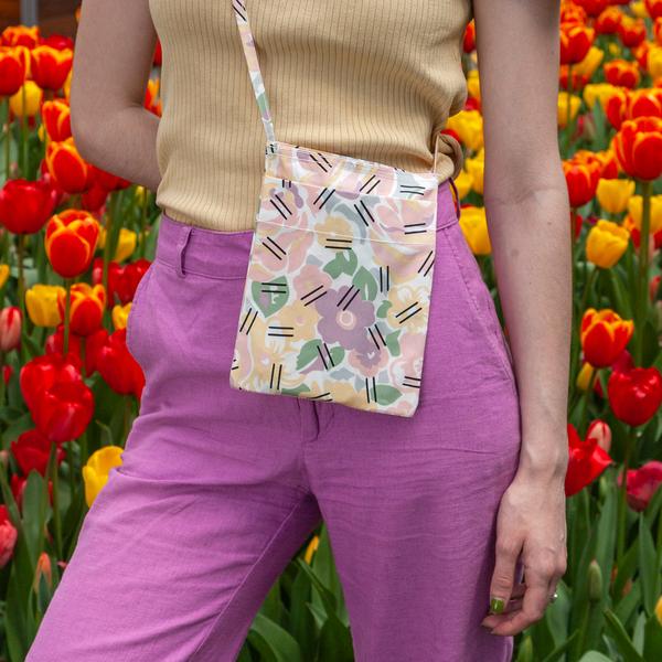 A model wears a small flat bag on her hip. The bag features light pastel-colored flowers with overlapping black hatch marks. The person is standing in a park with red and yellow tulips behind her.