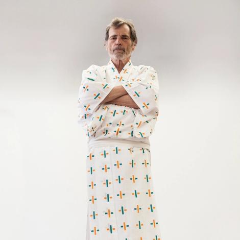 A photograph of a white man wearing a white kimono with criss-cross patterns in orange and green. He has graying hair and is staring directly at the viewer with his arms crossed in front of a white background.