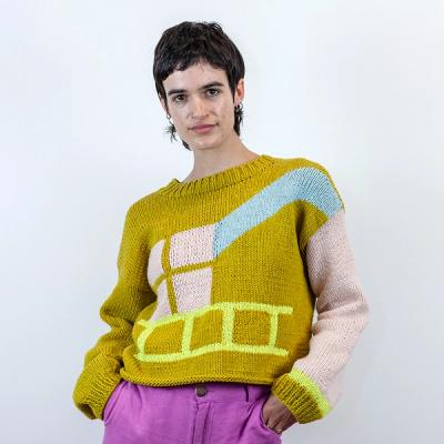 A person with short dark hair is wearing the sweater with pink pants. Their hands are in their pockets and they're looking at the camera while standing against a white background.
