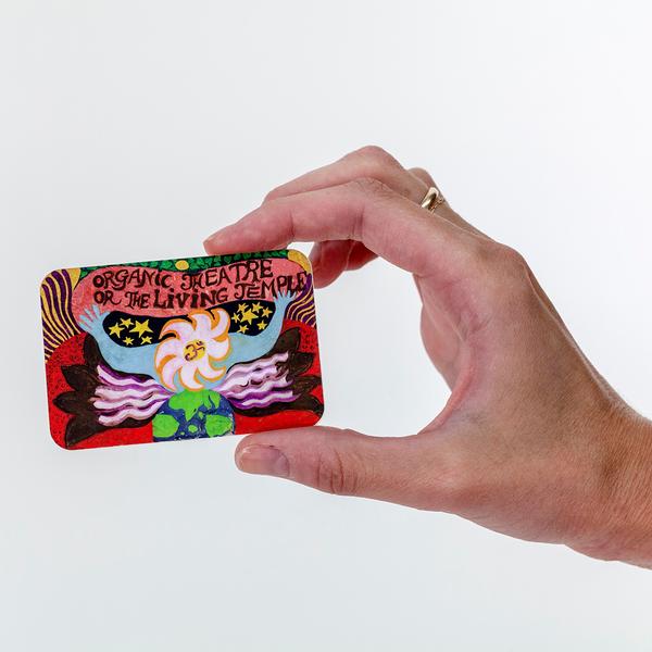 A hand holding out a colorful psychedelic sticker with bold text reading “Organic Theatre or The Living Temple” above a stylized figure with outstretched arms, swirling hair, and cosmic imagery of stars and the Earth.