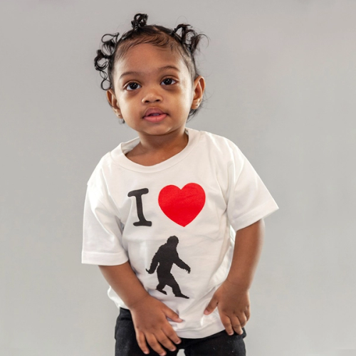A photo of an adorable Black child, around two years old, looking at someone slightly off-camera. She is wearing a white t-shirt with three characters: the letter I, the heart symbol, and a silhouette of the mythical creature, Bigfoot.