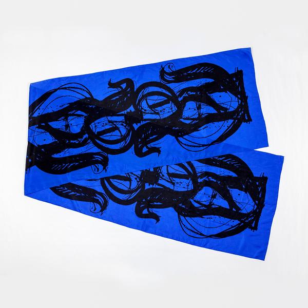 A silk scarf is folded and split diagonally against a white ground. This one is dark blue and features bold, painterly gestures with ink drips and splatters that follow the curving motion of a brush.