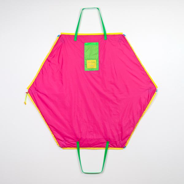 A hexagonally-shaped magenta bag hangs on a wall. It features two green straps, a green pocket, and yellow tag that reads "Risa Puno x FWM." The bag is outlined in yellow nylon.