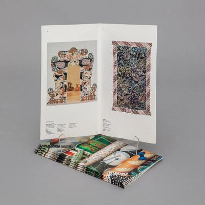 An image of a thin, rectangular book, more tall than it is wide. The book is open to show two artworks made of colorful, patterned fabrics.