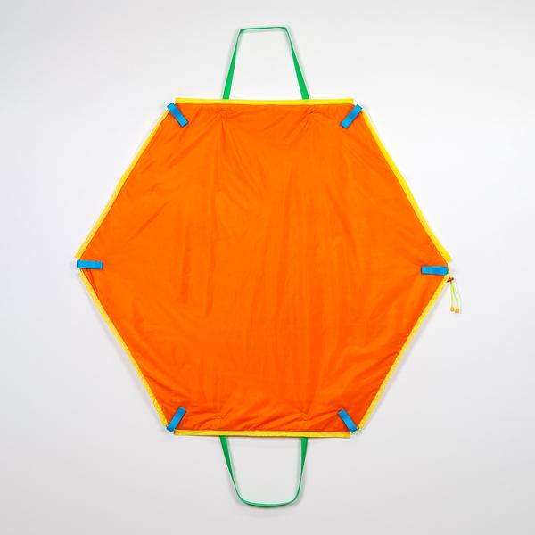 A hexagonally-shaped orange bag hangs on a wall. It features two green straps at the top and bottom and blue tabs on its corners. The bag is outlined in yellow nylon.