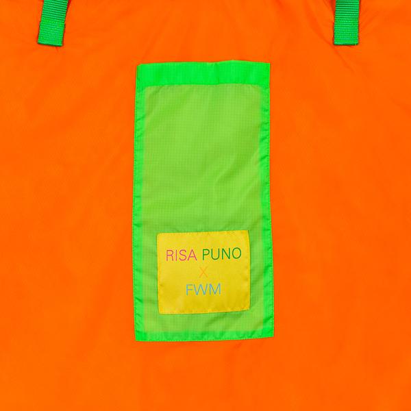 A detail of an orange bag with a green pocket and yellow tag that reads "Risa Puno x FWM." Green straps attach to the bag near the top of the image.