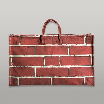 A frontal view of a broad handbag featuring a red brick pattern. Two sturdy handles form a loop on each side, secured in an ovular pinch over the brick pattern.