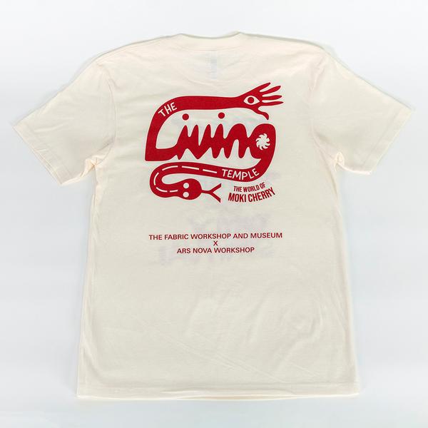 The back of an off white t-shirt featuring a red graphic logo that reads "The Living Temple: The World of Moki Cherry". The tail of the 'L' ends in a hand with an eye while the tail of the 'G' ends in a snake with a forked tongue protruding. The lower portion of the shirt reads "The Fabric Workshop and Museum x Ars Nova Workshop"