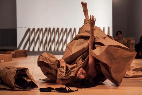 A woman performing in a gallery. She is laying on her back, with her right arm and right leg stretched upward towards the ceiling. She is wrapped up in several yards of brown packing paper. In the foreground you can see a black bra laying on the floor and off to the side is a large roll of brown packing paper. In the background you can see one guest watching the performance.