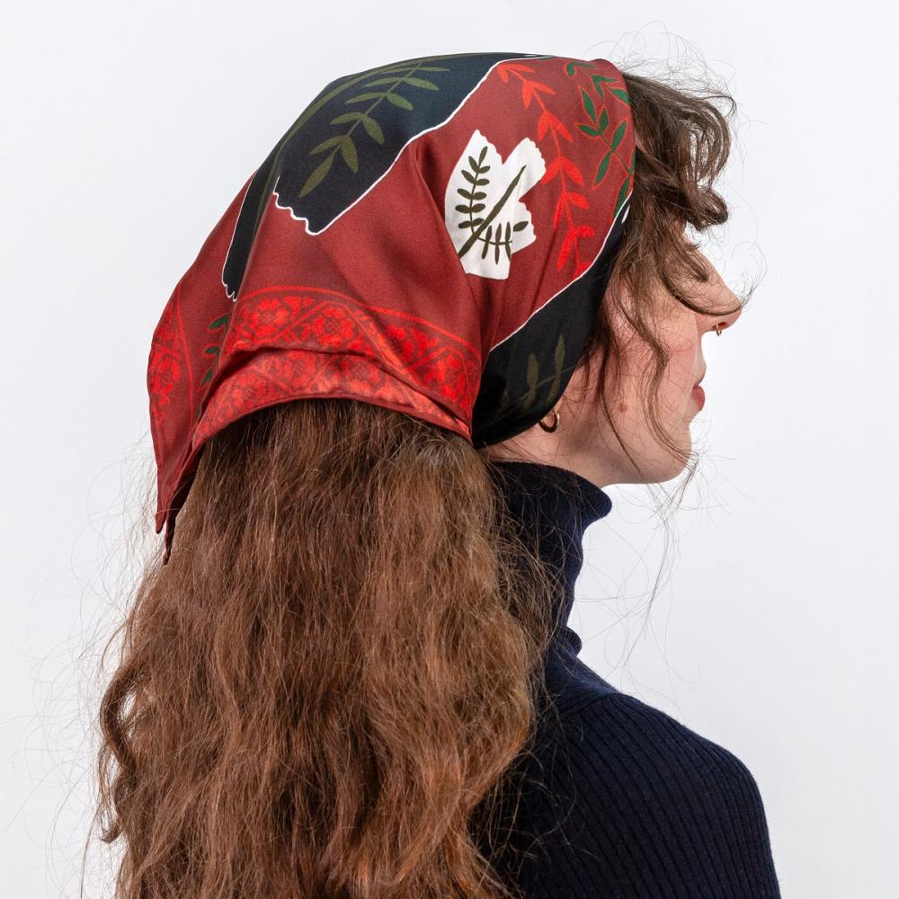 A portrait of the back side of a model wearing a red scarf tied atop her head. There is a large black dove illustration on the scarf sitting at the crown of her head with a white dove illustration below towards the side of her head. There is a floral motif bordering the edge of the scarf.
