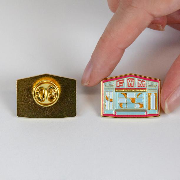An image showing the front and back of the pin side by side against a white backdrop. To the left, you see the back of the pin which is brass and has a butterfly clasp. To the right, you see the front of the pin being held by fingers coming from out of frame to show the scale.