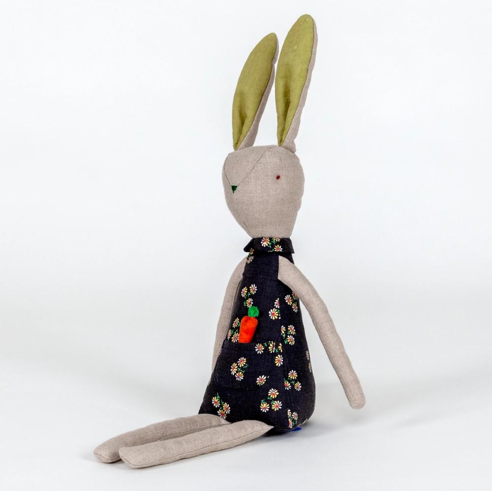 A photograph of a plush rabbit made of light beige woven fabric characterized to have a pointy nose, triangular shaped body, and long thin arms and legs. The body of the rabbit is made of a primarily blue patterned fabric and includes a pocket with a removable plush orange carrot.