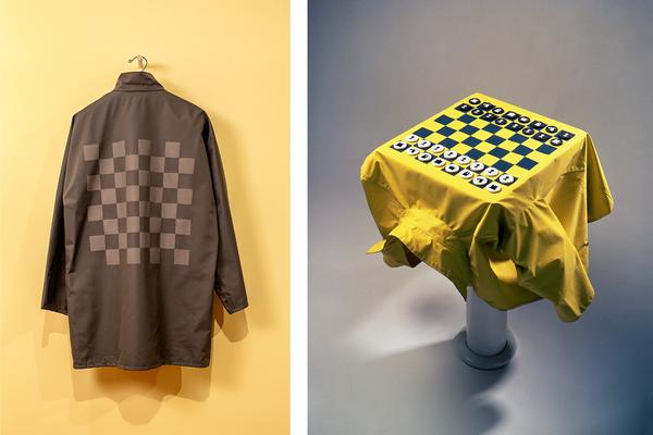 On the left, a gray-brown weatherproof jacket hangs from a hook against a pale yellow wall. The jacket features a chess checkerboard pattern printed in a lighter gray. On the right is a similar jacket, but in yellow, laid over a square table against a gray background. The checkerboard pattern, here printed in green, features small circular chess pieces in white and black arranged on each side, ready for a game of chess to begin.