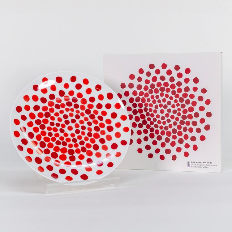 A photograph of a white plate with bold wobbly red dots in wobbly concentric circles. The dots are closer together in the center of the plate and become more spread out towards the edge. The dots do not vary greatly in size. To the right of the plate is a white box for the plate with the same red dots design.