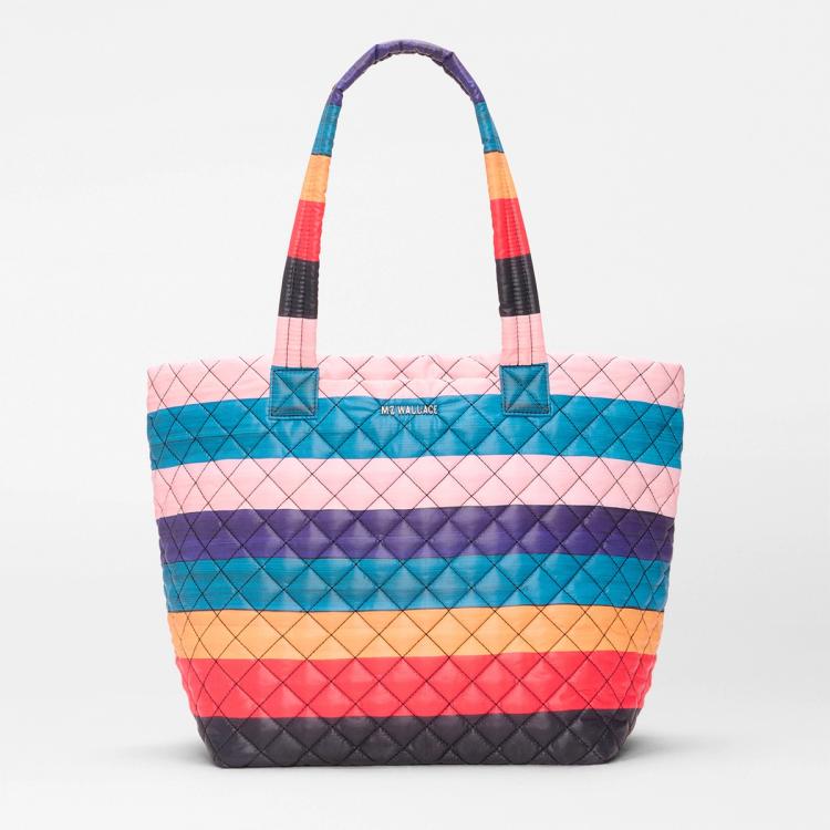 A photograph of a fabric quilted tote bag featuring painted horizontal stripes of alternating light pink, purple, blue, ochre, red, and black. The tote is sitting against a white backdrop.
