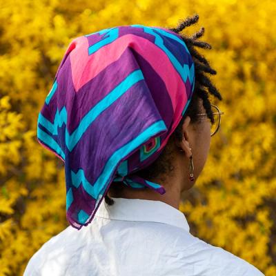 A young Black woman wearing a scarf tied to the back of her head stands in front of yellow foliage. She is facing away from the viewer. The scarf is mostly purple, with varying shades of blue and a bright pink border around the designs in the center.