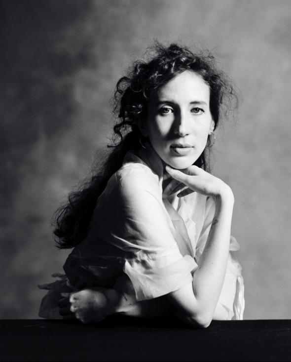 A black and white photo of a woman with curly dark hair sitting with her chin resting on her hand against a grey mottled backdrop.