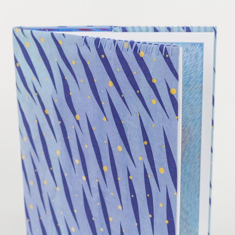 A photo of a hardback book with a hand-stitched book cover featuring a vibrant, pattern of diagonal dark blue brush-like streaks and scattered yellow dots on a light blue background. The book is slightly open, revealing a blue patterned illustration inside.