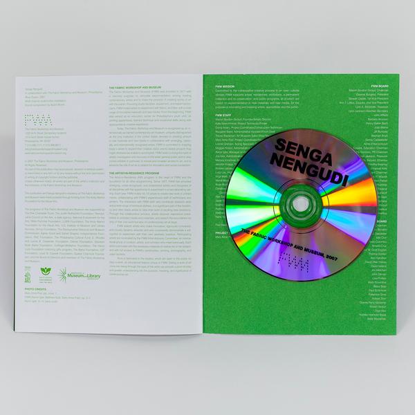 A photograph of an open book. On the left is a white page with green text featuring credit information for an exhibition. On the right the credit information continues on a green page with white text with a reflective CD positioned in the center.