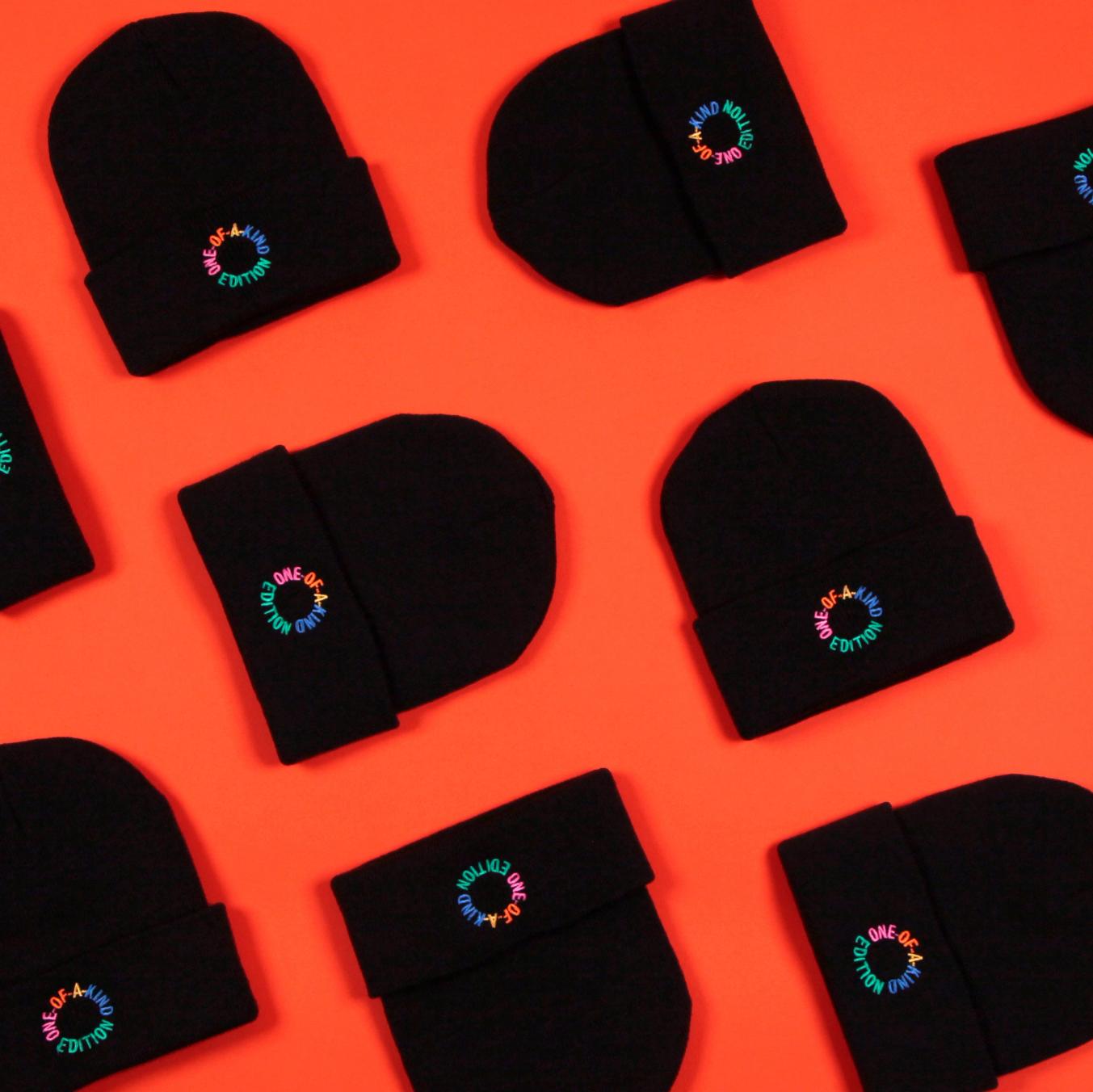 Multiple black beanies are set out in a grid against a tomato red background. Each beanie has a graphic that reads "one-of-a-kind edition" in a circular text layout. Each word is a different color, in pink, orange, yellow, blue, and green.