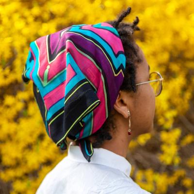 A young Black woman wearing a scarf tied to the back of her head stands in front of yellow foliage. She is facing away from the viewer. This scarf is primarily black, blue, and bright pink, with a purple border around the inner design and neon green accents.