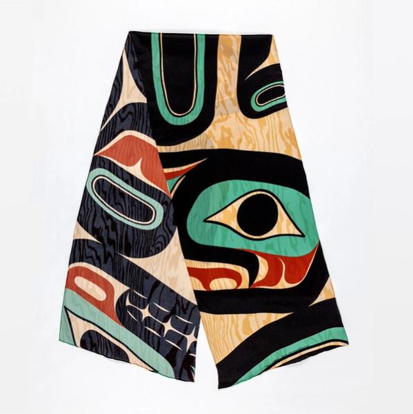 A scarf is folded and spread at a diagonal. The scarf has a texture mimicking woodgrain, and has a striking pattern in shades of tan, red, turquoise, and black.